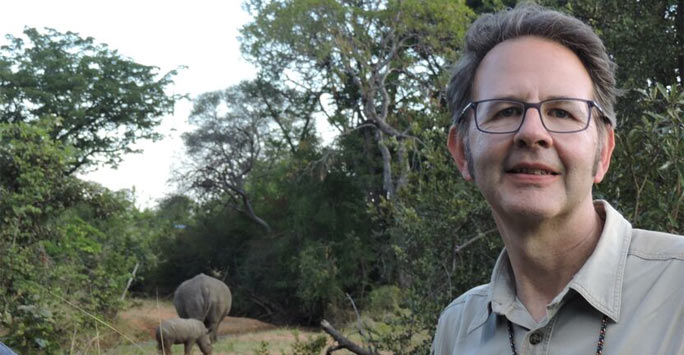 Professor Larry Barham smiles at the camera face on in a Zambian landscape, there are wild rhinos in the back of the picture