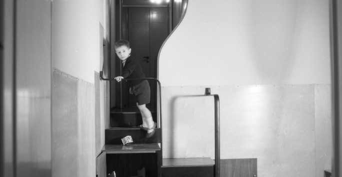 Male child wearing shorts standing halfway up a staircase, they have just dropped a cube with large letters on each face down the stairs.
