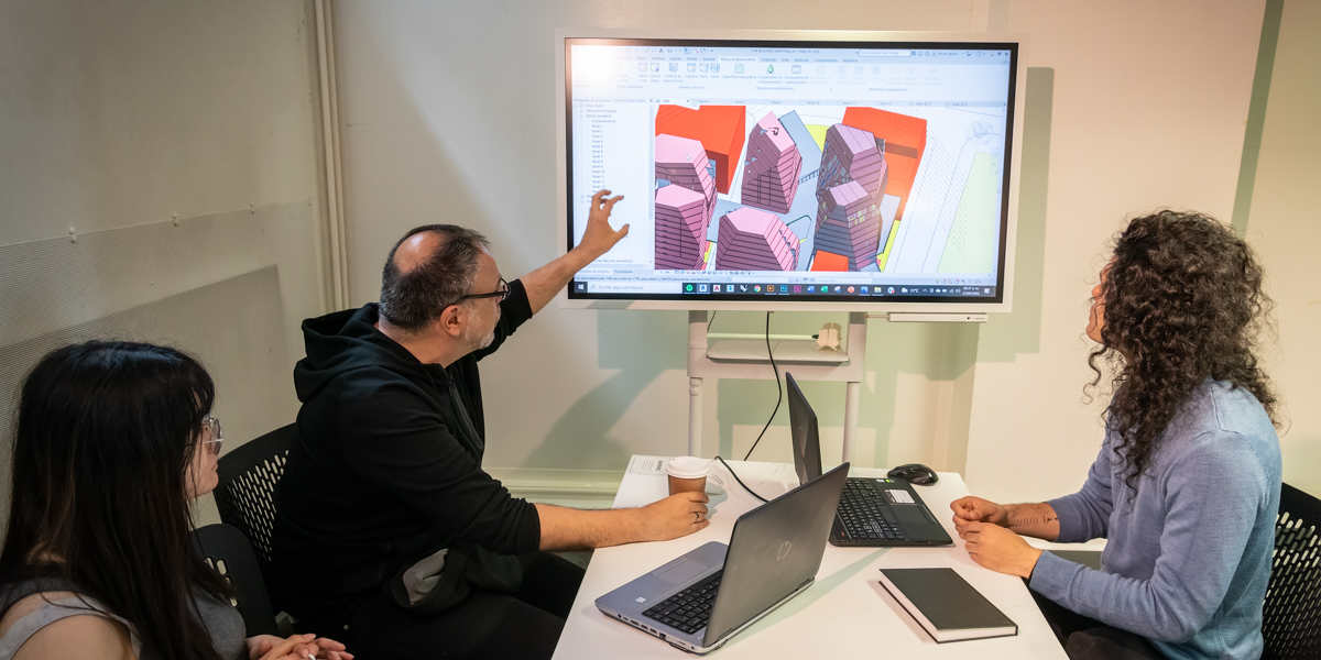 Thepeople sitting around a table looking at a large monitor didplaying an architectural scheme in 3D CAD programme. The one in the middle is pointing at the screen.