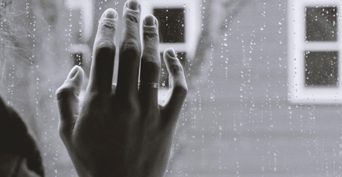 A black and white photo of a hand held against the inside of a rain soaked window