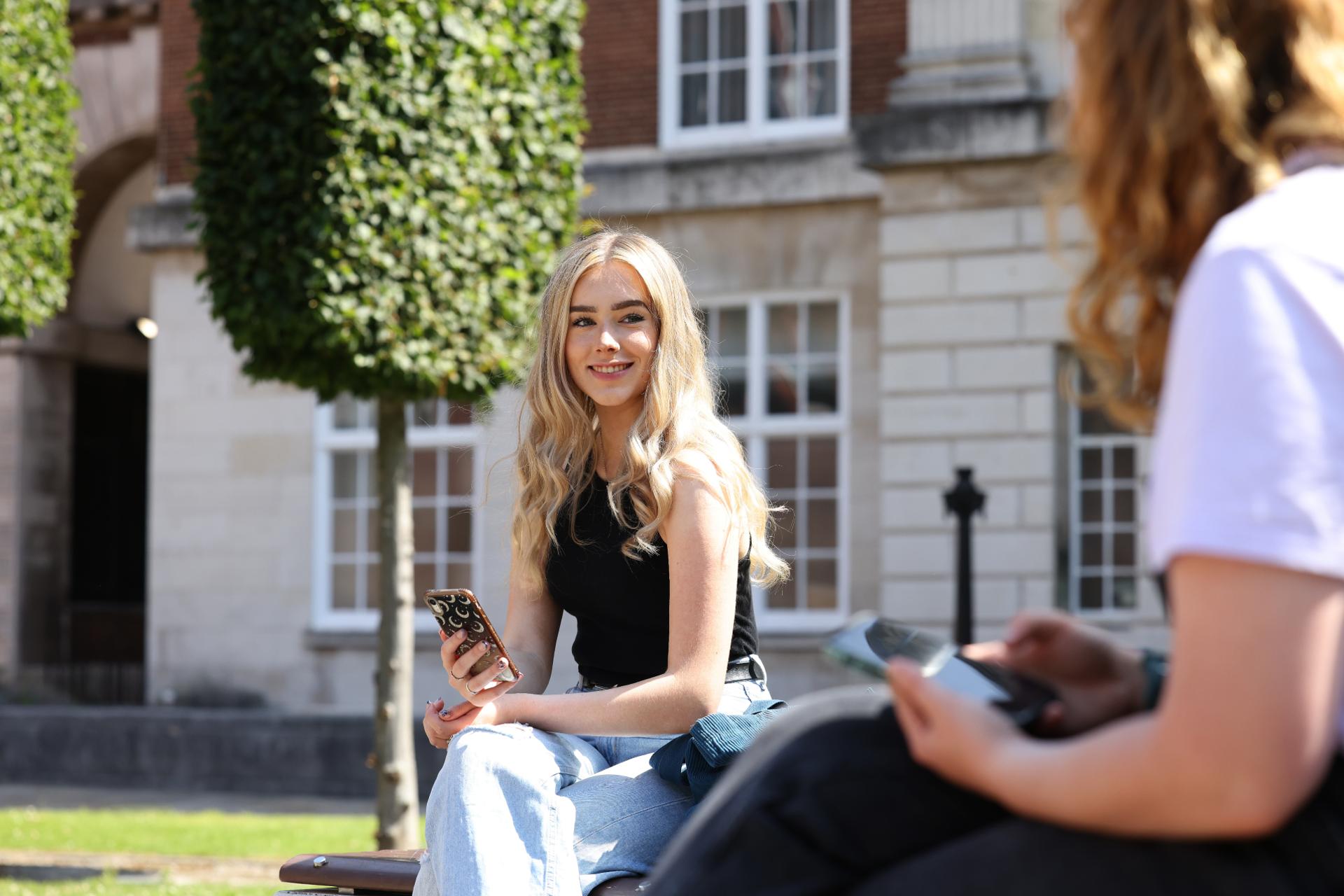 A student sat on a bench outside on campus, using a phone and smiling.