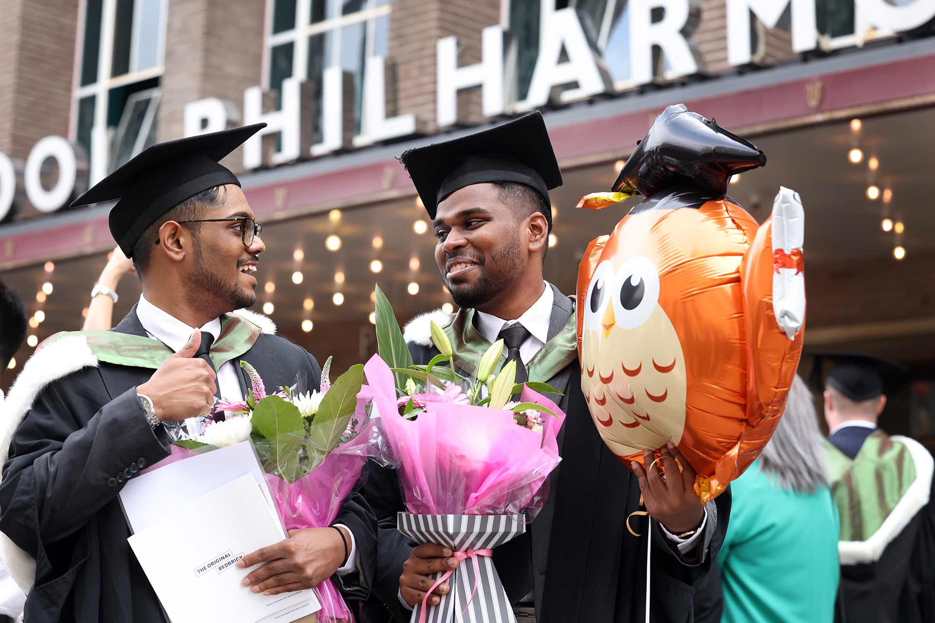Two graduates, holding a balloon and bouquet of flowers, standing outside the Liverpool Philharmonic Hall on their graduation day.