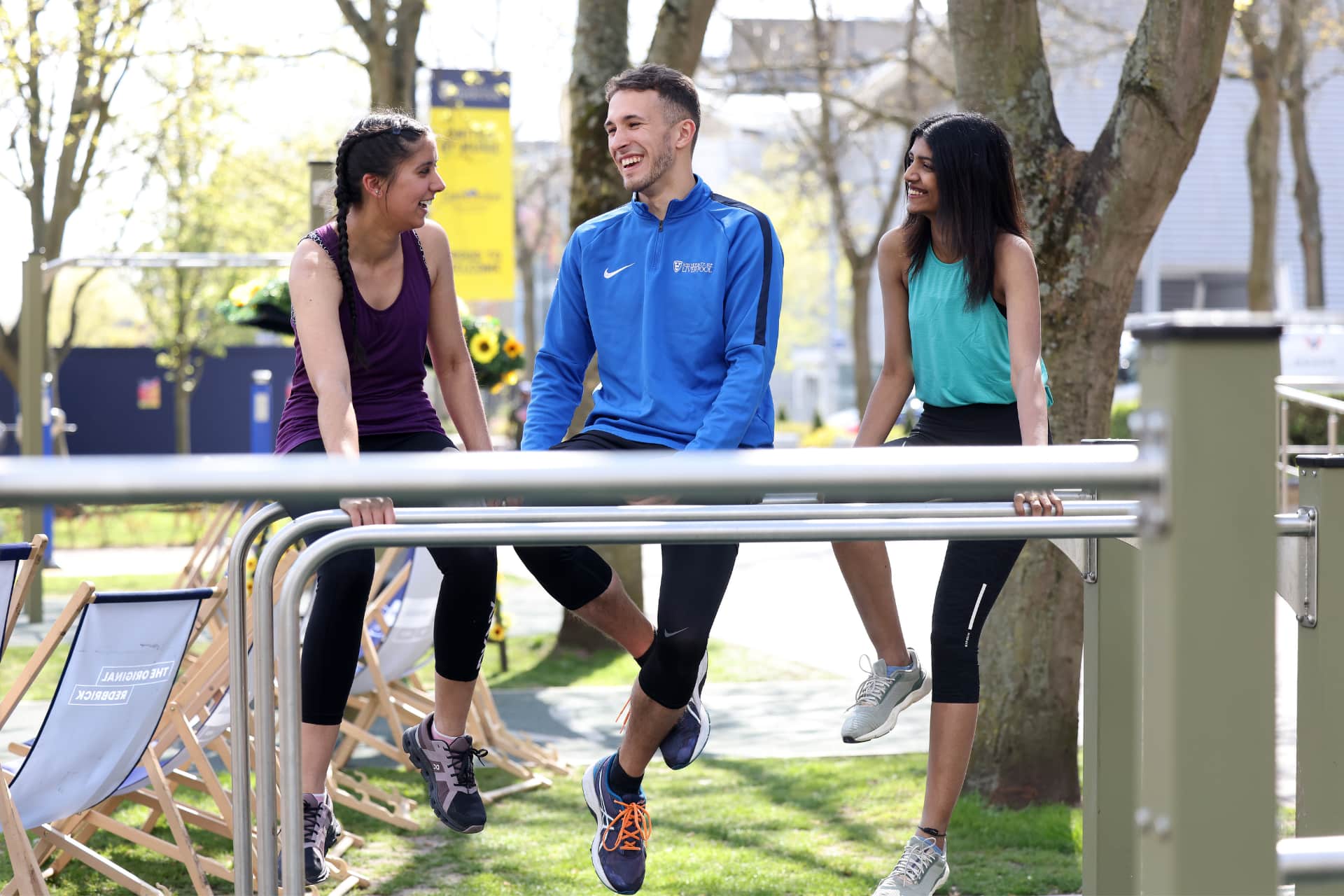 Three students sit on balance beams outside of the University of Liverpool's sport centre. They are wearing exercise clothes and smiling with each other.