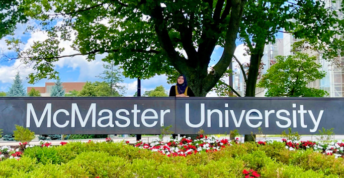 Sara Chowdhury standing behind the McMaster University sign, under a large tree with flowers to the foreground